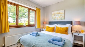 Una cama bien tendida con almohadas amarillas y azules, una manta verde y dos toallas enrolladas. Se ve una ventana con cortinas amarillas, un radiador y un cuadro en la pared.