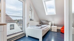 Ett sovrum med ett stort fönster, en säng, ett sängbord och en röd sittpuff.