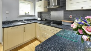 A modern kitchen with a granite countertop, beige cabinets, a stainless steel range hood, and a window with blinds. - 3 Bed in Abersoch (oc-sealav) (Abersoch)