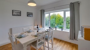 Un coin repas avec une table dressée pour quatre personnes, des chaises blanches, un radiateur et une fenêtre avec des rideaux.