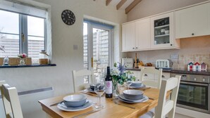 A dining area with a wooden table set for four, a clock on the wall, and a kitchen with white cabinets and a stove. - 2 Bed in Llanmadoc (oc-hh469) (Llanmadoc)