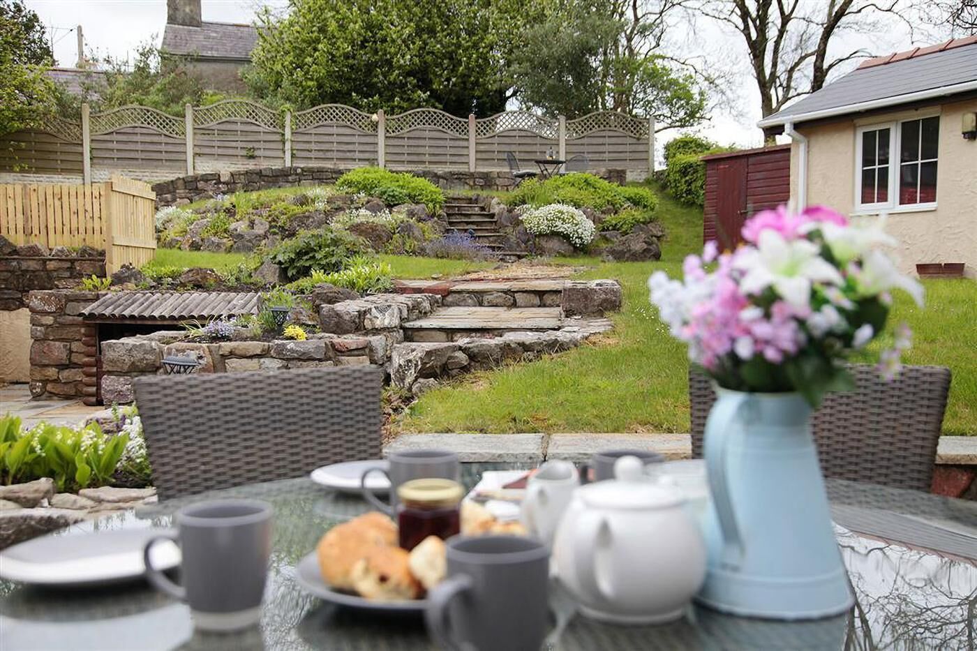 An outdoor dining area with a table set for a meal, surrounded by a stone wall and greenery.