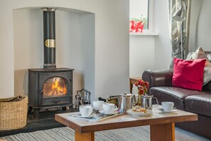 A cozy living room with a wood-burning stove, a leather sofa with a red pillow, and a coffee table with cups and a vase of flowers. - 3 Bed in Gower (oc-hh150) (Llangennith)