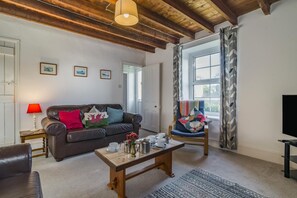 Een gezellige woonkamer met een leren bank, een houten salontafel en een blauwe fauteuil. Er hangen fotolijstjes aan de muur, een lamp op een bijzettafeltje en een raam met gordijnen.