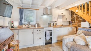 A compact kitchen with white cabinets, a stove, and a microwave. There's a wooden table with a chair, a small stool, and a television on the left. A sofa is in the foreground. - 1 Bed in Swansea (oc-hh404) (Llanmadoc)