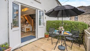 An open door leading to a cozy interior with a staircase, a dining area with a table and chairs, and a black umbrella providing shade. - 1 Bed in Swansea (oc-hh404) (Llanmadoc)