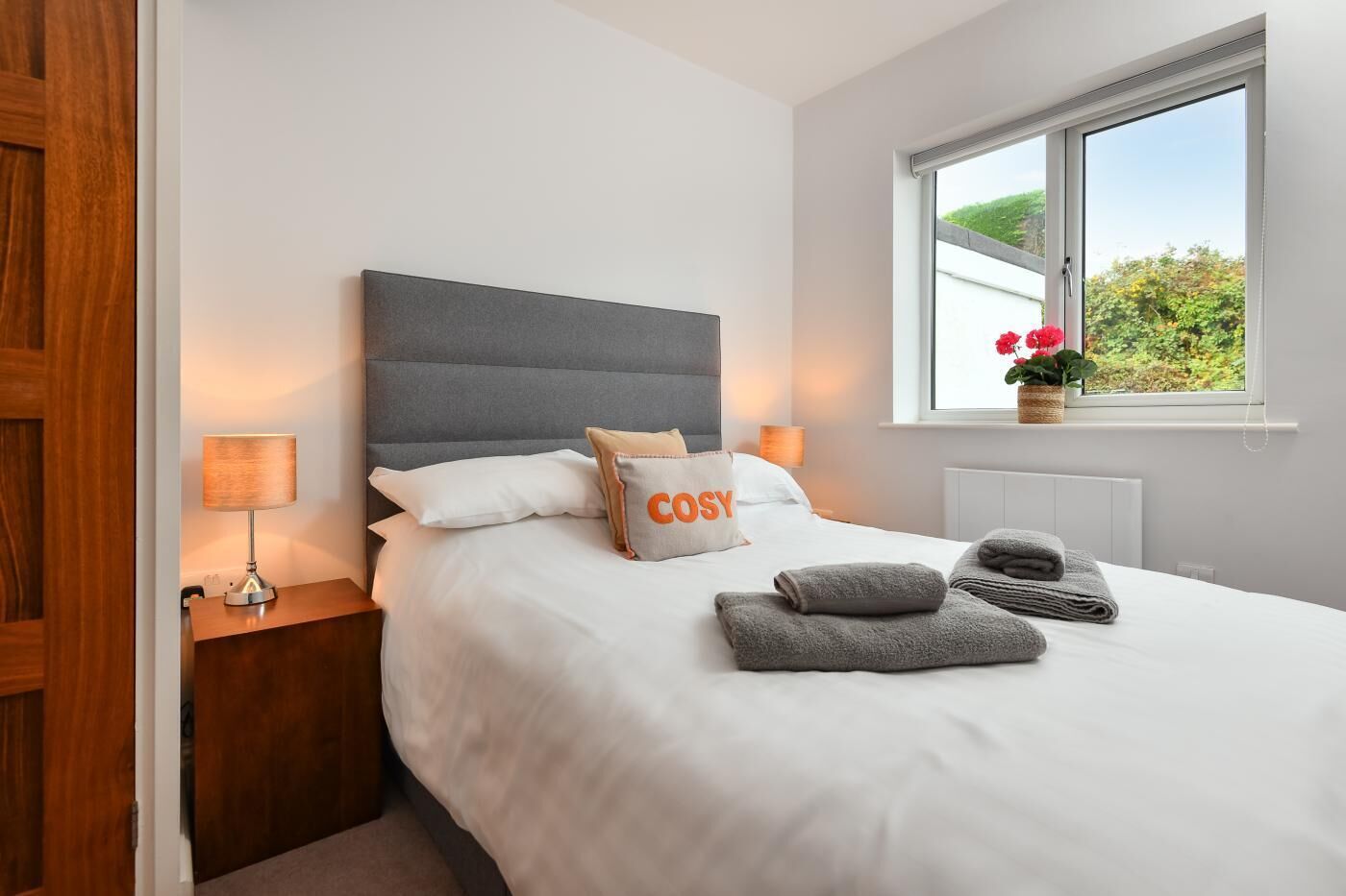 A neatly made bed with a grey headboard, white bedding, and a pillow with the word "COSTY". There are two bedside lamps, a window with a view of greenery, and a small potted plant on the windowsill.