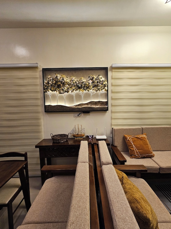 A living room with a sofa, a coffee table, a TV mounted on the wall, and a window with blinds.