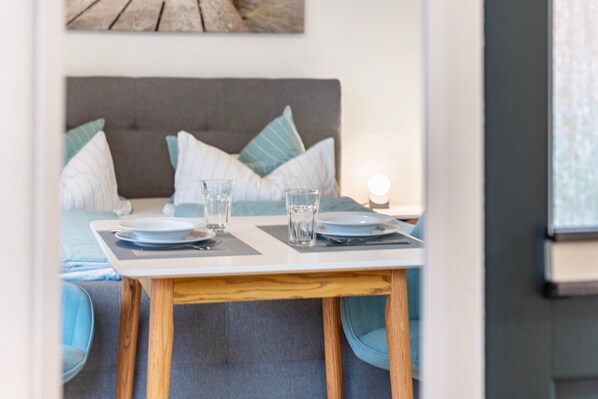 Une table à manger avec des assiettes et des verres, un canapé gris recouvert de tissu et, au fond, une tête de lit grise.