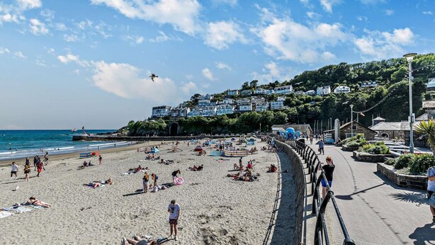 En strand där folk solar och badar, en strandpromenad med byggnader längs den och en klar himmel.