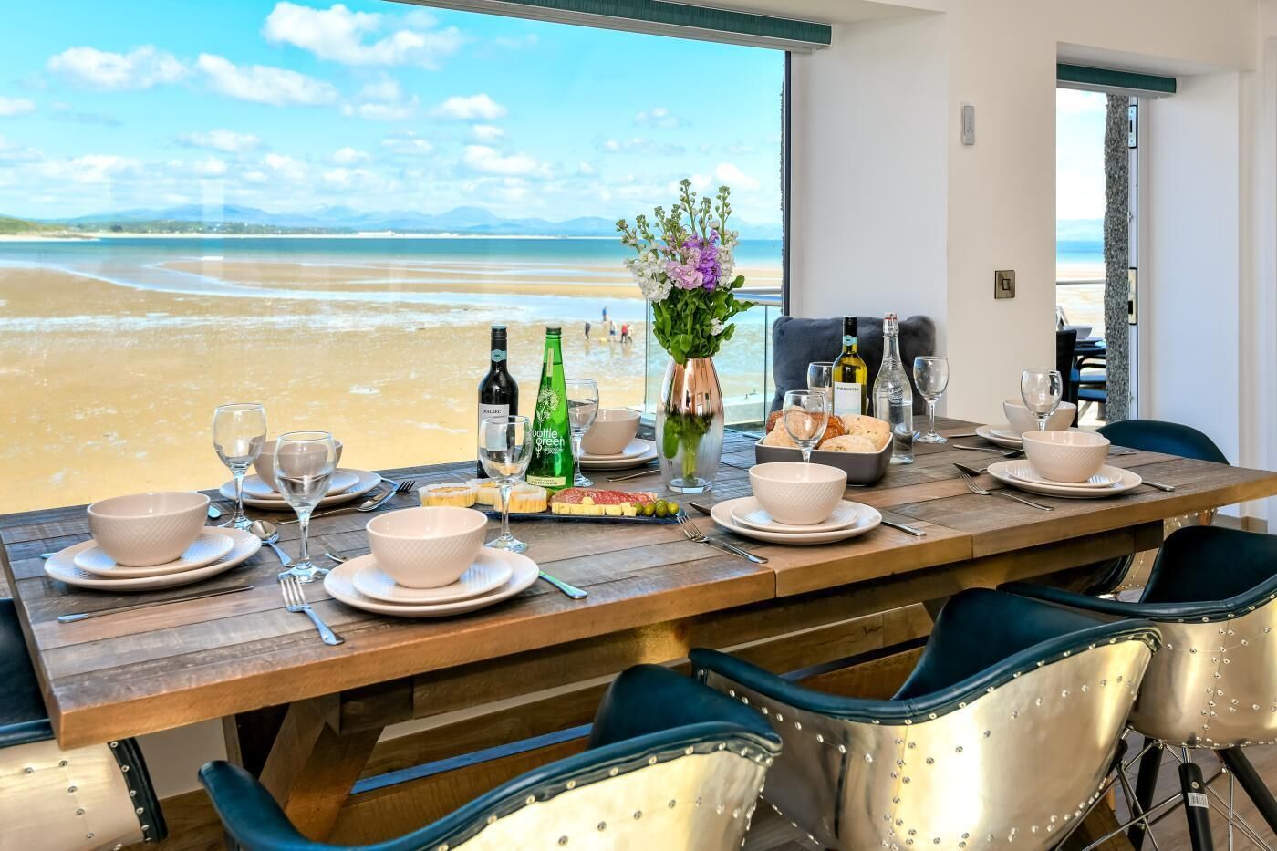A dining table set with plates, bowls, wine glasses, and bottles, overlooking a beach.