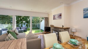 A modern living room with a sofa, dining table, and chairs. There is a fireplace, a television, and a balcony with outdoor furniture. - 2 Bed in Llanbedrog (oc-5glyny) (Llanbedrog)