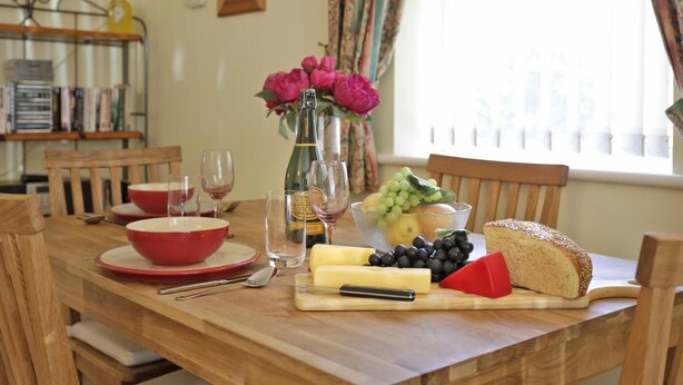 Une table de salle à manger en bois, avec un saladier rouge, des verres à vin, une bouteille de vin, un vase de fleurs, un panier de fruits, du fromage et du pain.