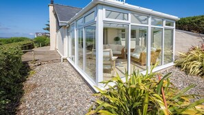 A modern glass house with a wooden deck, furnished with a sofa and a coffee table, surrounded by plants and a gravel path. - 2 Bed in Bwlchtocyn (oc-brynxx) (Bwlchtocyn)