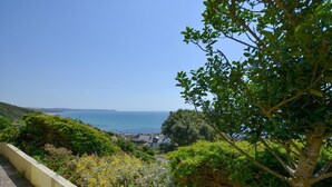 Paisagem costeira com vista para o mar, um morro com vegetação e um edifício ao longe.