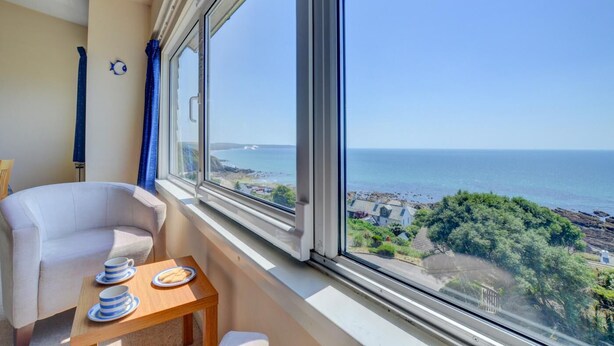Quarto com uma janela grande com vista para a costa, uma poltrona branca e uma mesinha com pratos e chávenas.
