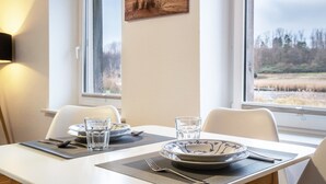Ein Esszimmertisch ist gedeckt mit Tellern, Gläsern und Besteck, ein Fenster bietet Blick auf eine Landschaft, und ein Bild hängt an der Wand.