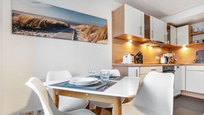 Eine moderne Küche mit weißen Schränken, einem Esstisch mit Stühlen und Wandbildern im Strandhausstil.