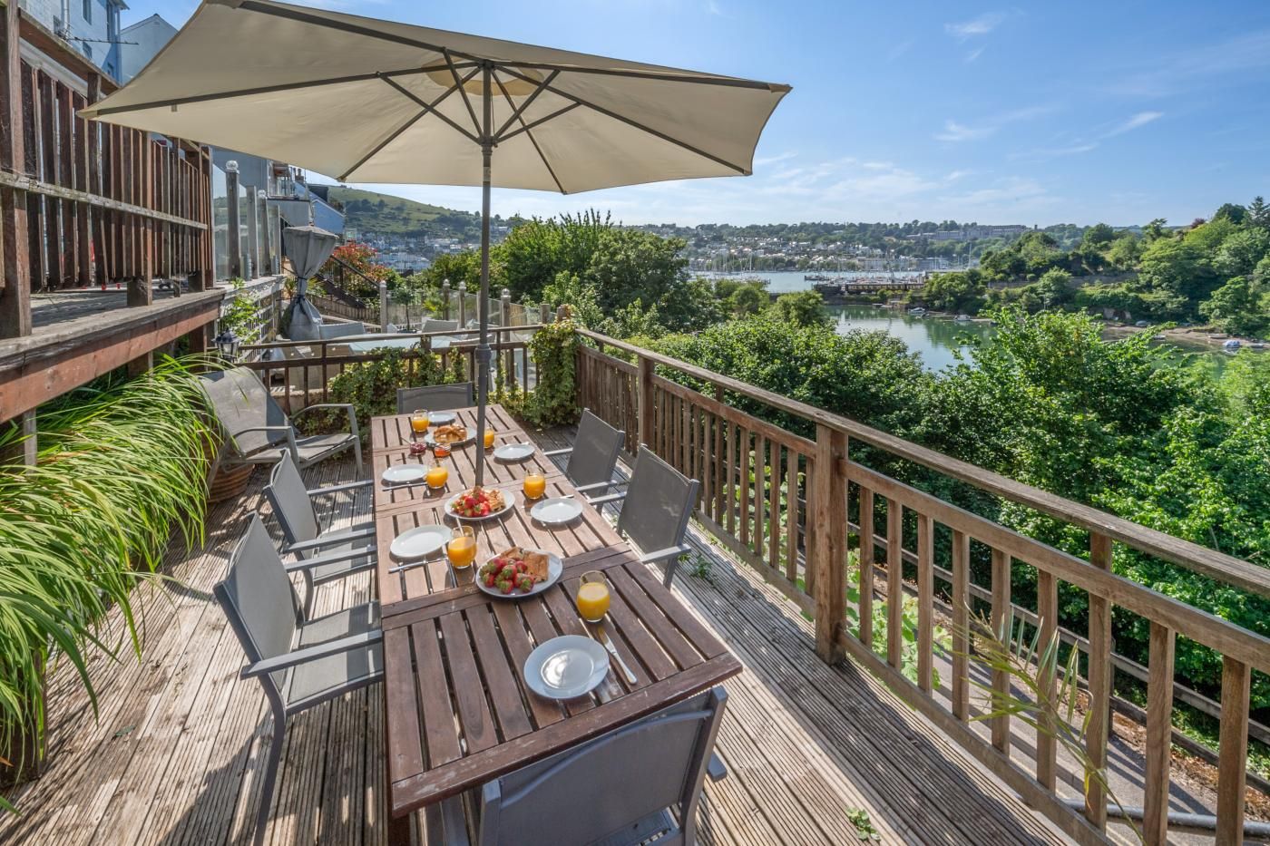 Una terraza de madera con una mesa preparada para el desayuno, con vistas a un río y una ciudad.