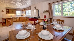 Une salle à manger avec une table en bois dressée pour six personnes, une cuisine en arrière-plan et une grande fenêtre avec des rideaux.