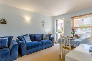 A living room with a blue sofa, a dining table with chairs, a wall clock, and a window with blinds. - 2 Bed in Aldeburgh (oc-7cc) (Aldeburgh)