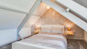 Ett mysigt sovrum på vinden med ett sluttande tak, en säng med en vit sänggavel, två sänglampor och en tapet med mönster.