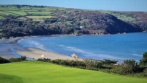 Een golfbaan aan de kust met een zandstrand, een vlag op de green en glooiende heuvels op de achtergrond.