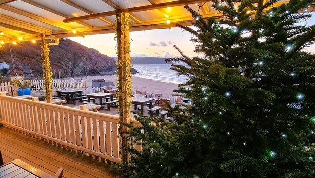 Een terras aan het strand met tafels en stoelen, een witte picket fence en een kerstboom met lichtjes.
