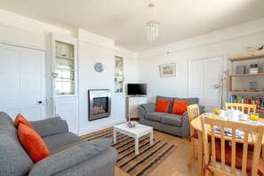 A living room with a fireplace, a television, a dining area with a table and chairs, and a bookshelf. - 2 Bed in Southwold (oc-5eas) (Southwold)