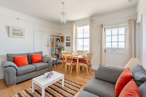 A living room with a grey sofa, a white coffee table, a dining table with chairs, a bookshelf, and a window with curtains. - 2 Bed in Southwold (oc-5eas) (Southwold)