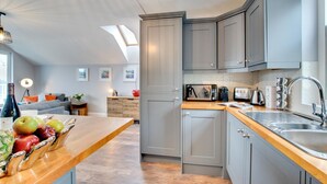 Eine moderne Küche mit grauen Schränken, einer Holzarbeitsplatte und einem eingebauten Backofen. Es gibt einen Essbereich mit einem Tisch für zwei Personen und einen Blick auf das Wohnzimmer.