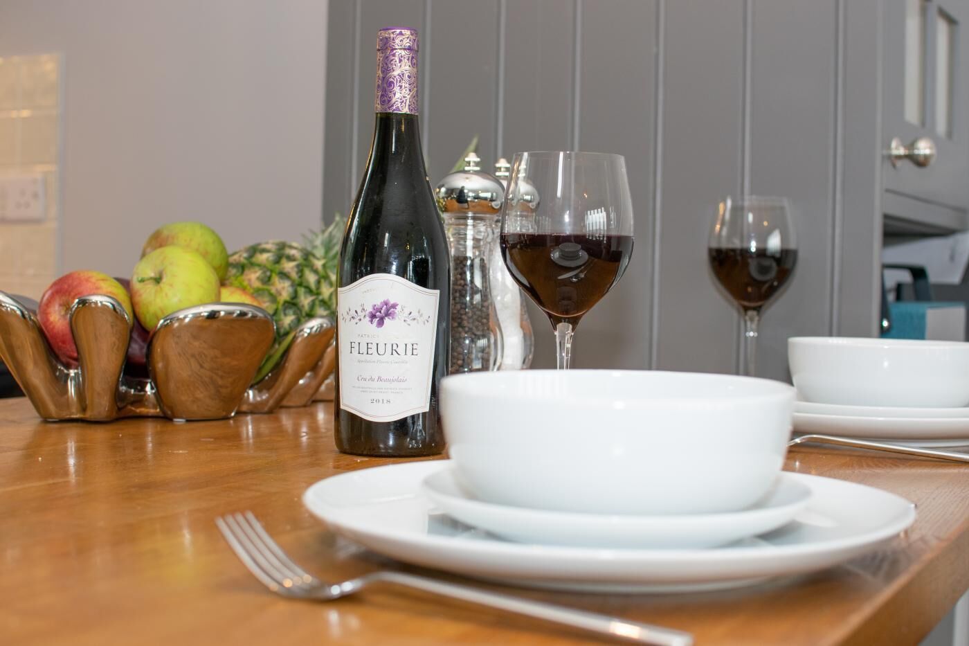 A table with a bottle of Fleurie wine, two glasses of red wine, a bowl, and a fruit bowl with apples.