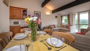 A dining area with a table set for four, a vase of flowers, and a kitchen in the background. - 3 Bed in Llanbedrog (oc-rosell) (Llanbedrog)