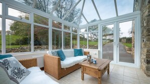 A conservatory with a wicker sofa, a wooden coffee table, and a view of the garden. - 4 Bed in Llanbedrog (oc-bragdy) (Llanbedrog)