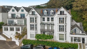 A multi-story residential building with a central balcony, a staircase, and a parking area with several cars. - 2 Bed in Fowey (oc-p00839) (Fowey)