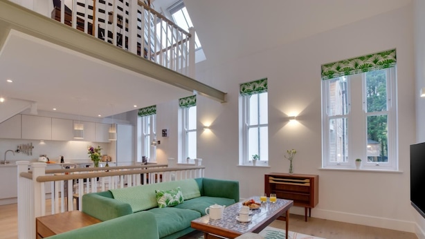 A spacious living area with a green sofa, a coffee table, and a TV. There is a kitchen in the background with white cabinets and a dining area. - 2 Bed in Fowey (oc-p00703) (Fowey)