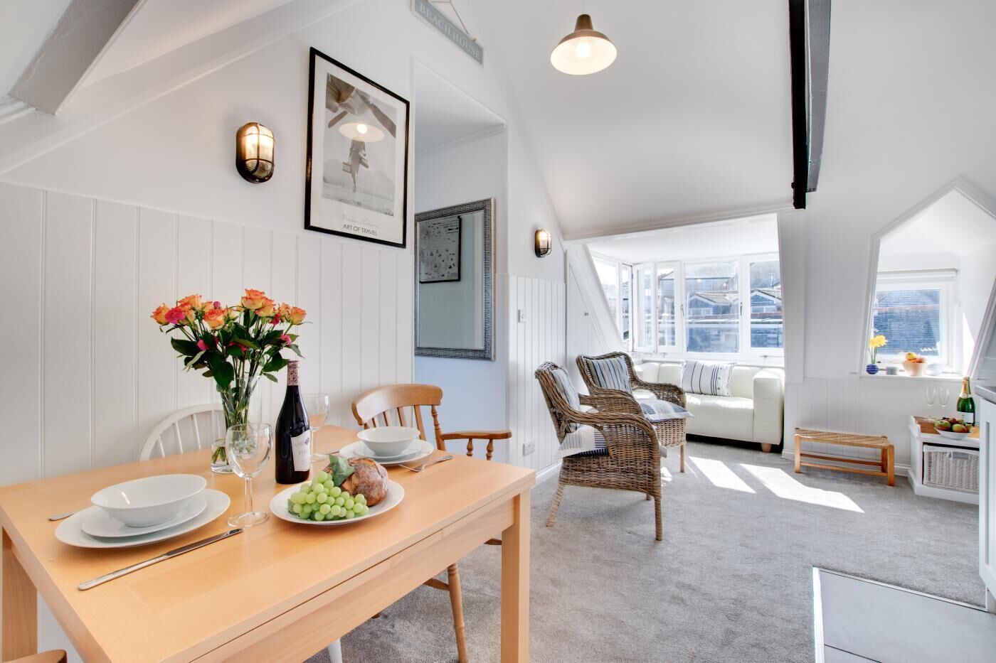 Un coin repas avec une table en bois dressée pour deux personnes, un vase de fleurs et une bouteille de vin. On aperçoit en arrière-plan des chaises en osier et un canapé.