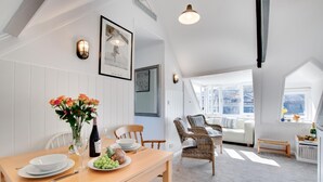 Ein Essbereich mit einem Holztisch, gedeckt für zwei Personen, einer Blumenvase und einer Weinflasche. Im Hintergrund sind Korbstühle und ein Sofa zu sehen.