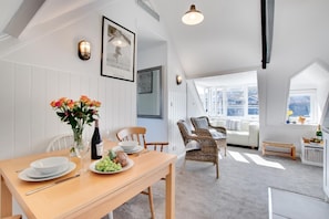 A dining area with a wooden table set for two, a vase of flowers, and a bottle of wine. There are wicker chairs and a sofa in the background. - 1 Bed in St. Ives (oc-cp5) (St Ives)