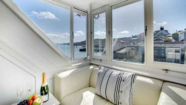 Ein Zimmer mit einem weißen Sofa, einem kleinen Tisch mit Flasche und Gläsern sowie Blick auf einen Hafen mit Gebäuden und einer Brücke.