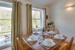 Uma sala de jantar com mesa de madeira posta para seis pessoas, uma garrafa de vinho e vista para o exterior pela janela.