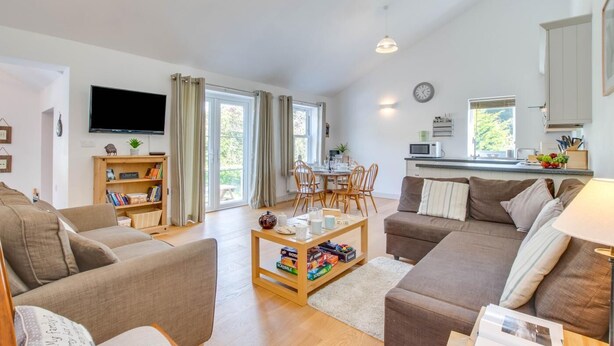 A modern living room with a sofa, a coffee table, and a bookshelf. There is a television mounted on the wall and a dining area with chairs and a table. - 2 Bed in Llaniestyn (oc-gweger) (Llaniestyn)