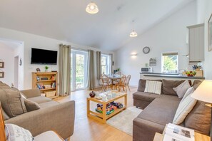 A modern living room with a sofa, a coffee table, and a bookshelf. There is a television mounted on the wall and a dining area with chairs and a table. - 2 Bed in Llaniestyn (oc-gweger) (Llaniestyn)