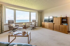 Ein Hotelzimmer mit großem Fenster, zwei Sesseln, einem Couchtisch, einem Fernseher und einem Bücherregal.