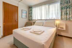 Ein ordentlich bezogenes Bett mit weißen Leinen und Handtüchern, eine Holztür, ein Stuhl und ein Fenster mit durchsichtigen Vorhängen.