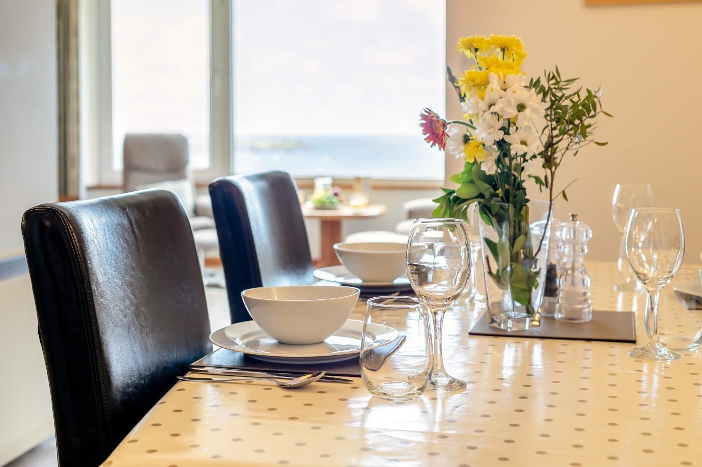 A dining table set with plates, bowls, and wine glasses, with a vase of flowers and a view of the outdoors.