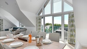 Ein Essbereich mit Tisch für vier Personen, umgeben von Korbstühlen, einem großen Fenster mit Vorhängen und Blick auf Grünfläche.
