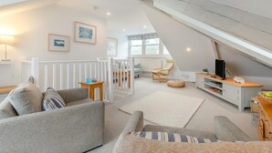 Une pièce de style « loft », avec un plafond haut, une rambarde blanche, un canapé, un fauteuil, une petite table, un téléviseur et une fenêtre.