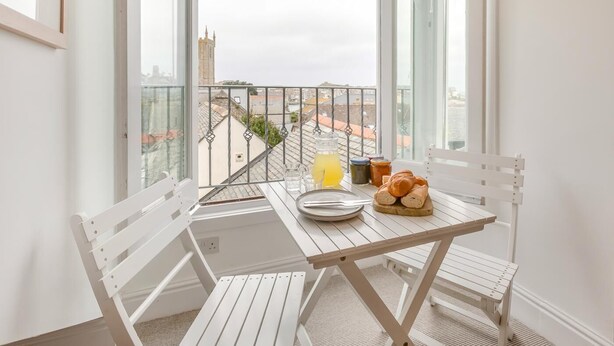Un balcon avec une table, des chaises, un verre de jus de fruits, du pain et des pots de confiture.