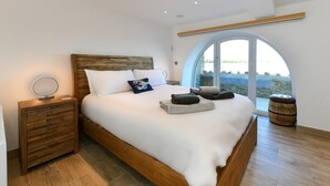 Um quarto espaçoso com uma cama grande, cabeceira de madeira e vista através de uma ampla janela em arco.